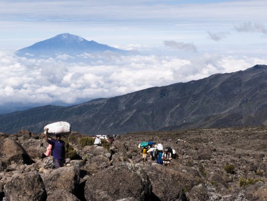 Mt Kilimanjaro, Tanzania, East Africa - February 11th 2010: 8.45am - A long trail of porters carrying provisions and equipment up along the the Machame Route up Mt Kilimanjaro having just left Shira Camp. The peak of Mount Meru breaking cloud cover in the background.