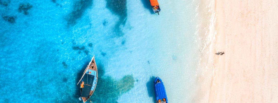 Aerial view of the fishing boats in clear blue water at sunset in summer. Top view from of boat, yacht, sea and walking man on the sandy beach in Zanzibar. Travel. Tropical seascape. View from above